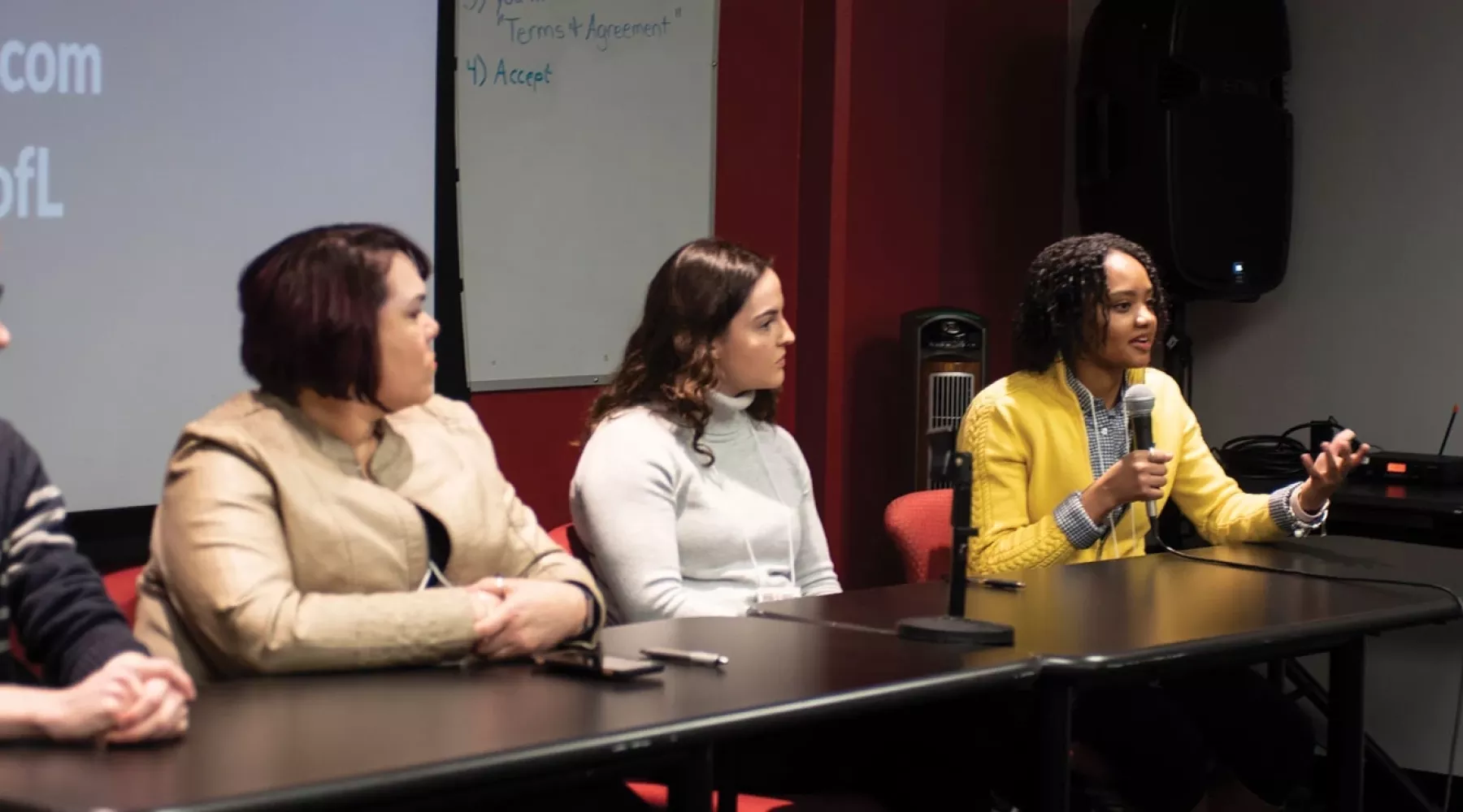 People sitting at a table with one person using microphone to speak to audience during a presentation