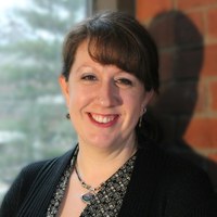 A woman wearing a cardigan over a patterned shirt standing in front of a brick wall. 