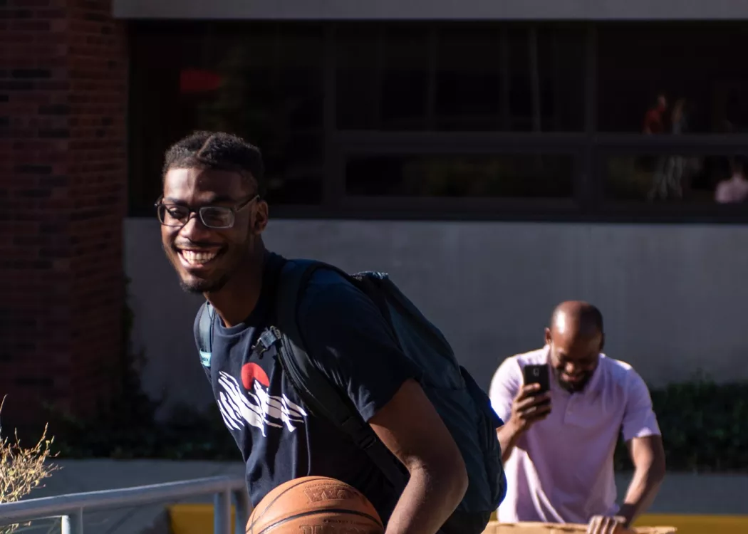 Smiling student rolls a suitcase up the sidewalk to his dorm housing.