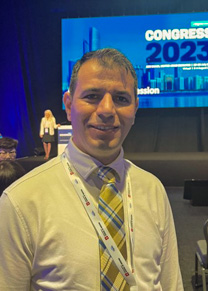 A smiling man in a white button up shirt and a blue yellow and white plaid tie stands with a presentation on a large screen behind