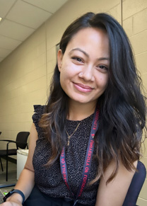 A smiling woman in a short sleeved dark top is seated in an office 