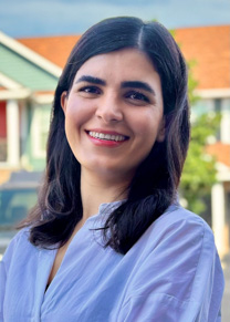 A smiling woman with shoulder length dark hair stands outside in front of a building 