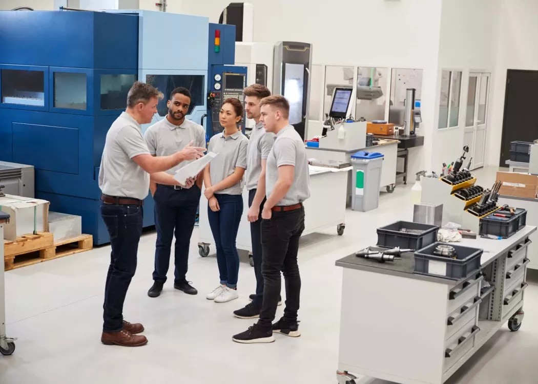 Engineering Team Meeting On Factory Floor Of Busy Workshop